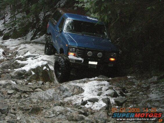 Blue Ford Bronco with large tires and auxiliary lights on a rocky, snowy trail.