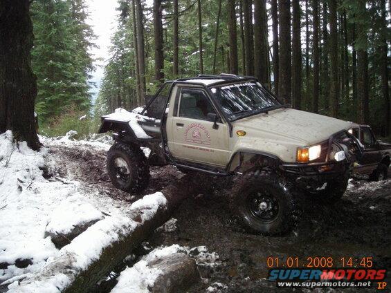 Modified tan Toyota pickup truck off-roading in a snowy, muddy forest.