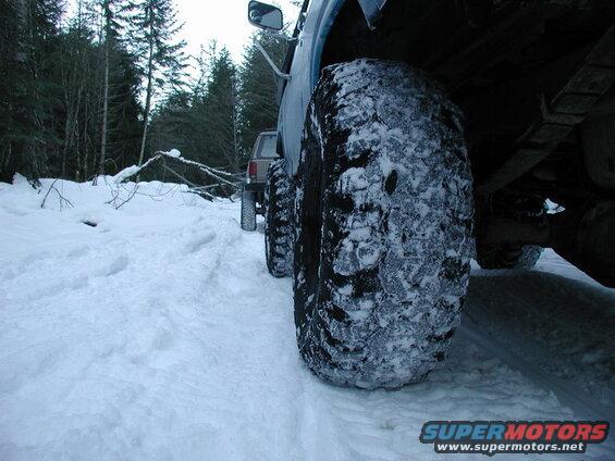 Close-up of a snow-covered tire on a blue Ford Bronco in a snowy forest.