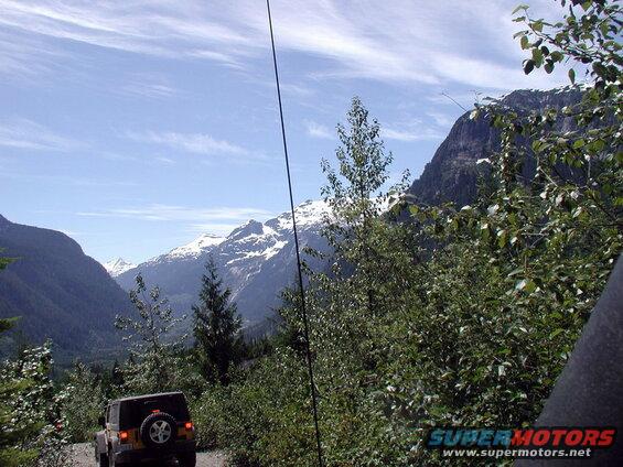 Yellow Jeep Wrangler driving on a mountain road with snow-capped peaks in the background.