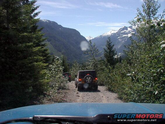View from inside a blue vehicle on a dirt road with two Jeep Wranglers ahead, mountains in the distance.