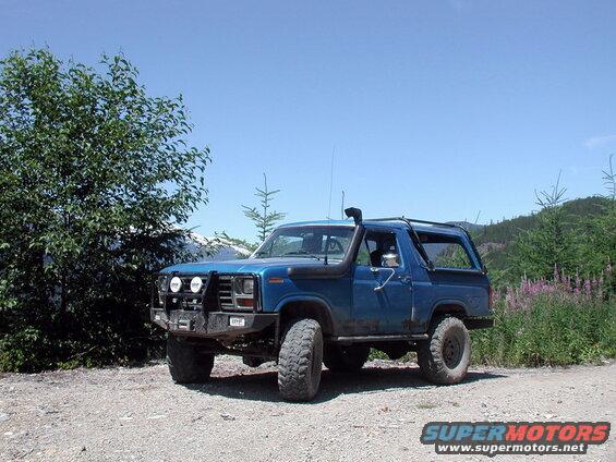 Blue 1980s Ford Bronco with off-road mods parked on gravel road, trees and mountains in background.
