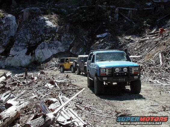 Blue 1980s Ford Bronco with brush guard and snorkel on a debris-strewn dirt road.