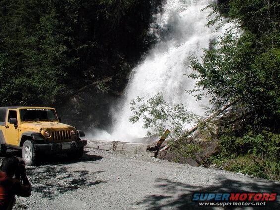 Yellow Jeep Wrangler Rubicon parked on gravel road by a waterfall.