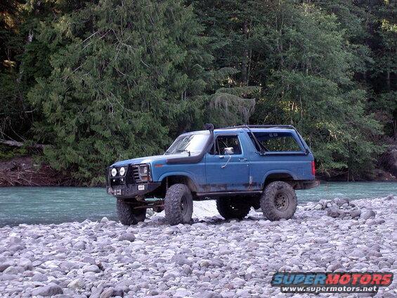 Blue Ford Bronco parked on a rocky riverbed with a forest and river behind it.