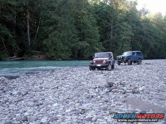 Maroon Jeep Wrangler and blue Ford Bronco on a rocky riverbank with a turquoise river and forest.