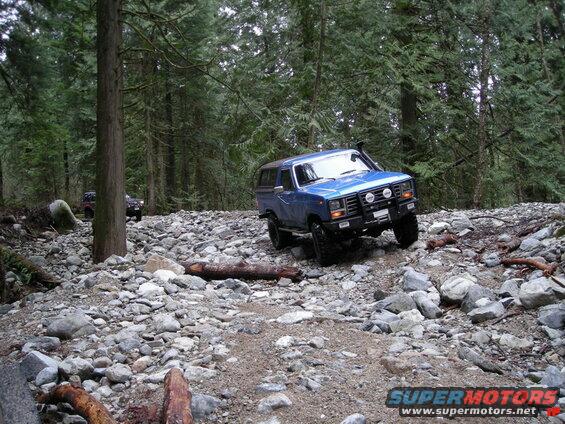 Blue 1980s Ford Bronco off-roading on a rocky trail in a forest.