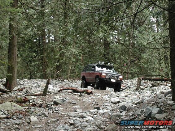 Red SUV driving on a rocky forest trail with roof-mounted lights