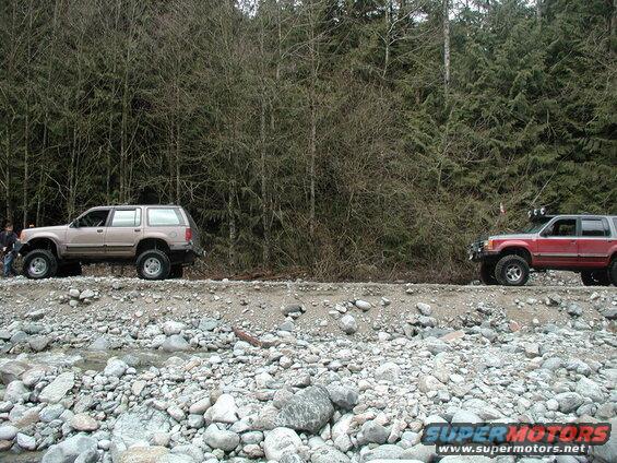 Two lifted SUVs parked on a rocky embankment with a forest behind them.