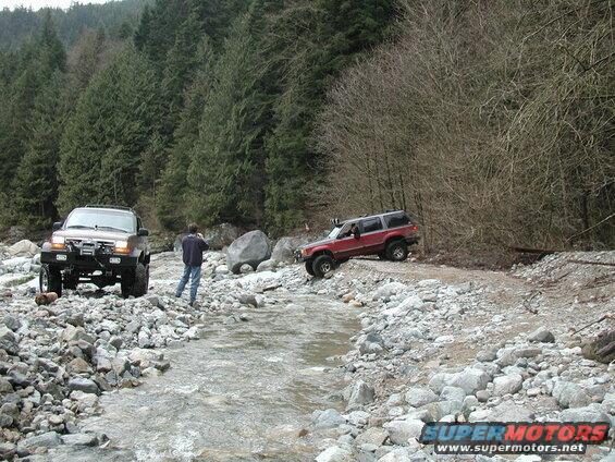 Two off-road SUVs in a rocky riverbed with a forest backdrop.