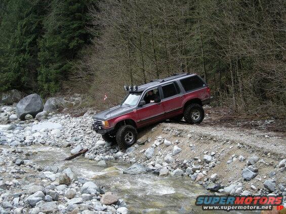 Red and gray SUV driving on a steep, rocky bank next to a stream.