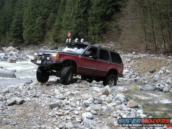 Lifted red SUV driving through a rocky river with a forest backdrop.