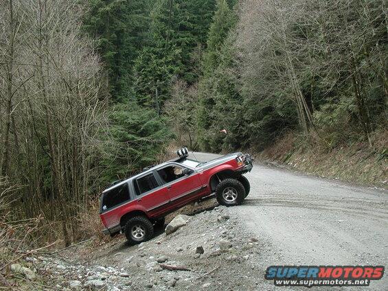 Red SUV tilted on a steep, loose gravel embankment next to a dirt road in a forest.