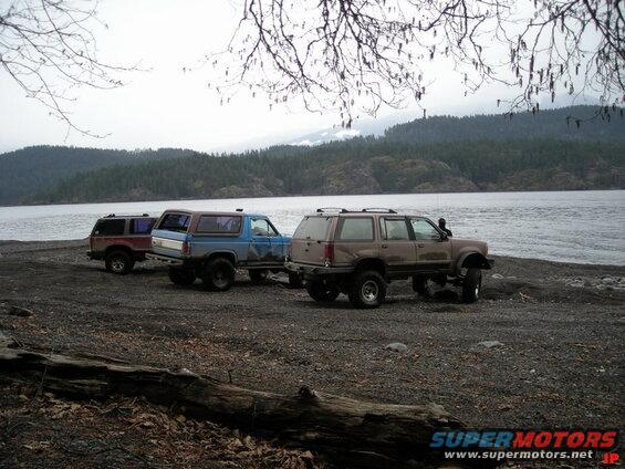 Three modified SUVs parked on a rocky shore by the water.