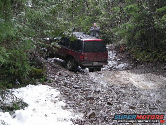 Red SUV on a muddy and rocky forest trail with a man on its roof.