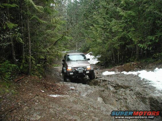 Tan SUV driving on a muddy forest road with snow patches.