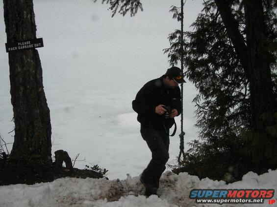 Person in snow holding a camera next to a tree with a "Please Pack Garbage Out" sign.