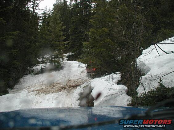 View from inside a blue vehicle driving up a snowy forest trail with another vehicle ahead.