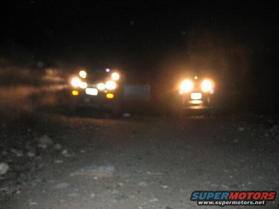 Two blurry vehicles at night on a dirt road with headlights on.