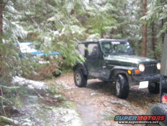 Green Jeep Wrangler in a snowy forest with another vehicle in the foreground.