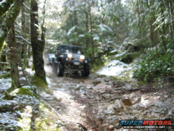 A Jeep Wrangler drives on a muddy, snowy forest road.