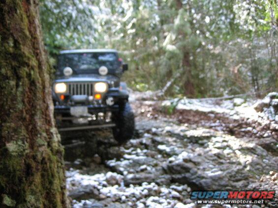 Dark Jeep Wrangler driving on a snowy, rocky forest trail.