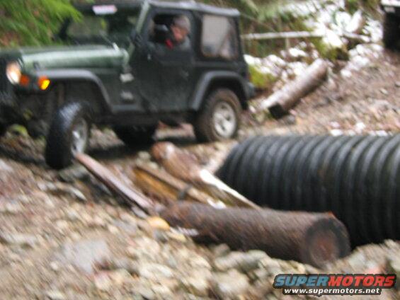 Green Jeep Wrangler driving on a muddy trail with logs and a culvert.