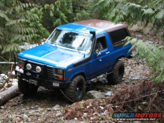 Blue 1980s Ford Bronco off-roading on a rocky, wooded trail.