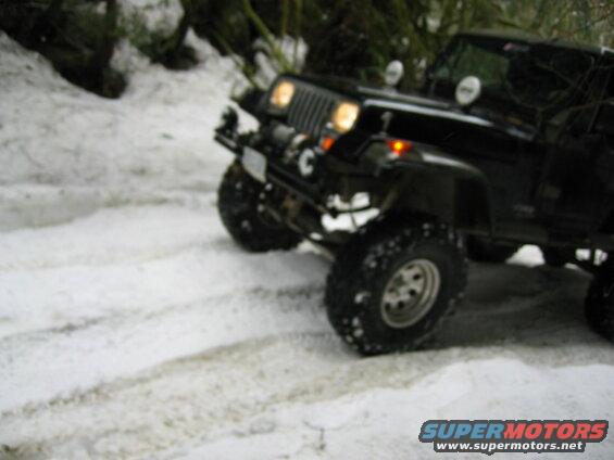 Black Jeep Wrangler with off-road tires and winch driving on a snowy road.