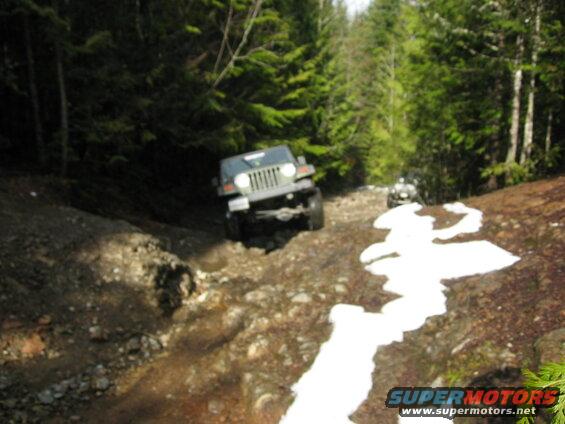 Jeep Wrangler driving up a steep, rocky, wooded trail with snow.
