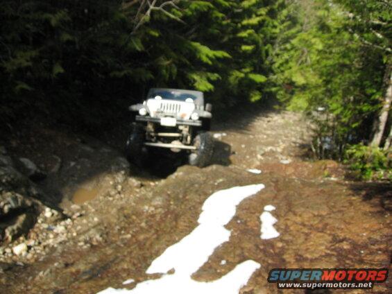 White Jeep Wrangler driving on a muddy, rocky trail through a forest.