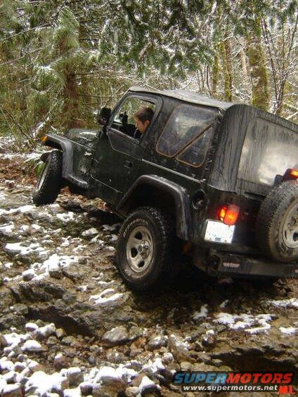 Dark green Jeep Wrangler driving on a rocky, snow-dusted forest trail.