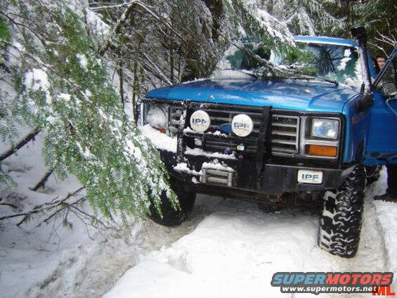 Blue 1980s Ford Bronco in a snowy forest, partially covered by a tree.