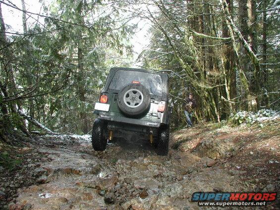 Dark green Jeep Wrangler driving on a rocky, muddy forest trail with snow.