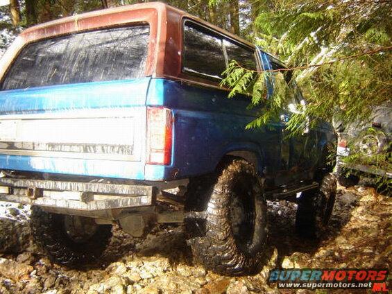 Rear passenger side of a blue Ford Bronco with a brown roof on a rocky, wooded trail.