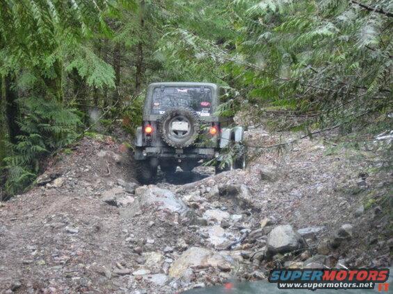Jeep Wrangler driving on a rocky, muddy off-road trail in a forest.