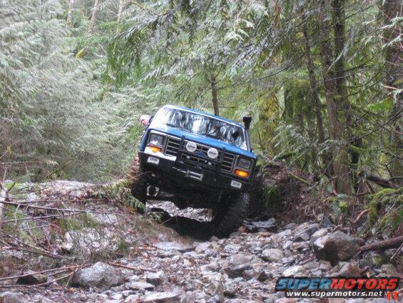 Blue 1985 Ford Bronco off-roading on a rocky, wooded trail.