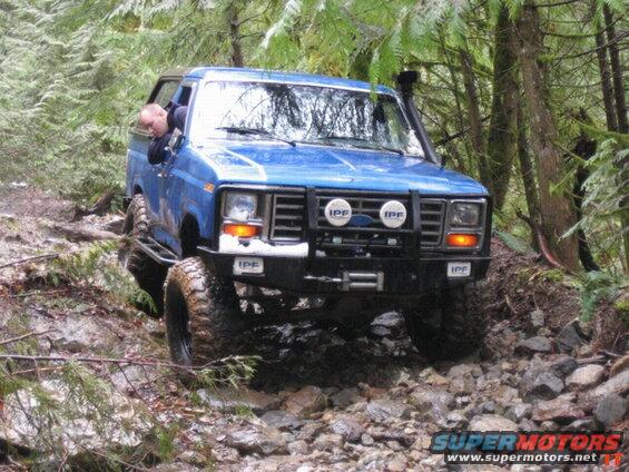 Blue 1980s Ford Bronco off-roading on a rocky, wooded trail.