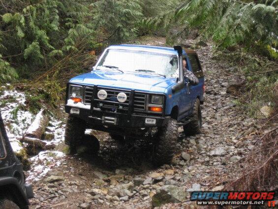 Blue 1980s Ford Bronco on a rocky, wooded trail with snow.