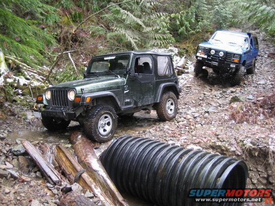 Green Jeep Wrangler and blue Ford Bronco off-roading on a muddy, wooded trail.