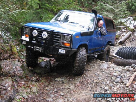 Blue 1980s Ford Bronco off-roading on a rocky, wooded trail.