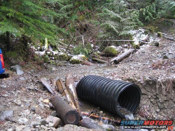 Blue vehicle rear and a large pipe discharging water in a wooded, rocky area.