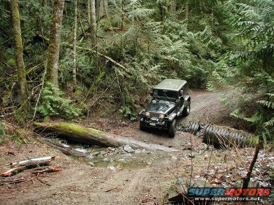 Dark green Jeep Wrangler on a muddy forest road crossing a stream.
