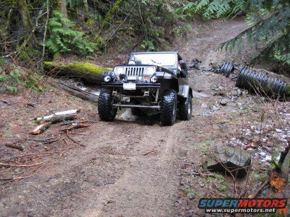 Dark Jeep Wrangler on a muddy forest trail with large tires.