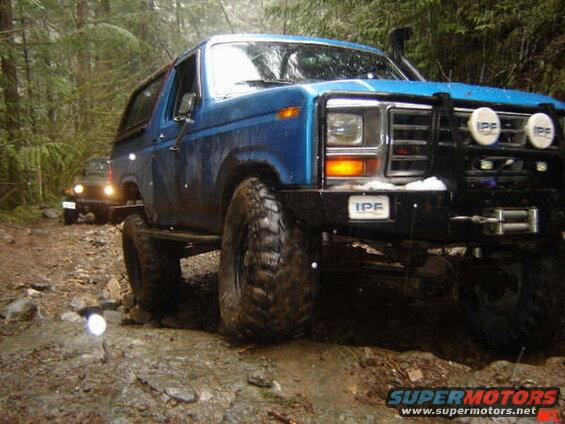 Blue Ford Bronco off-roading on a muddy, rocky forest trail.