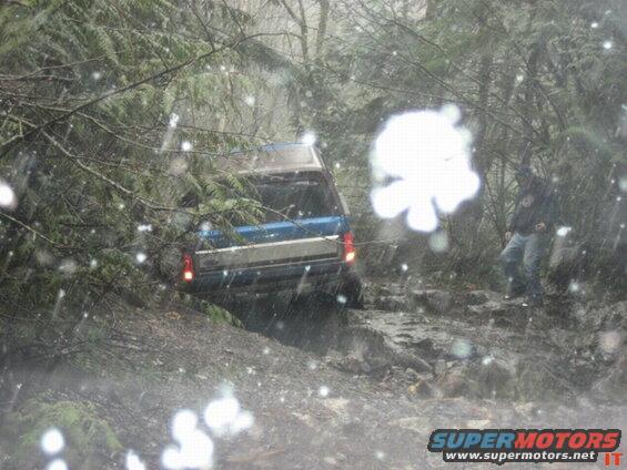 Blue and silver Ford Bronco on a muddy, rocky forest trail, viewed from rear.