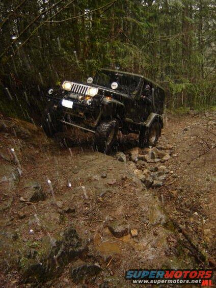 Black Jeep Wrangler driving on a wet, rocky forest trail.