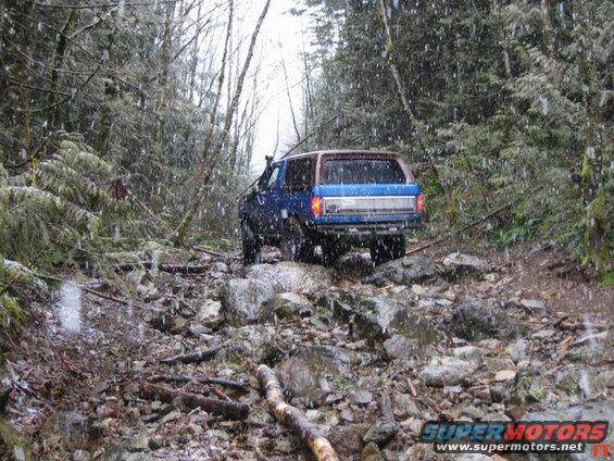 Blue 1980s Ford Bronco driving on a rocky, wooded trail in the snow.