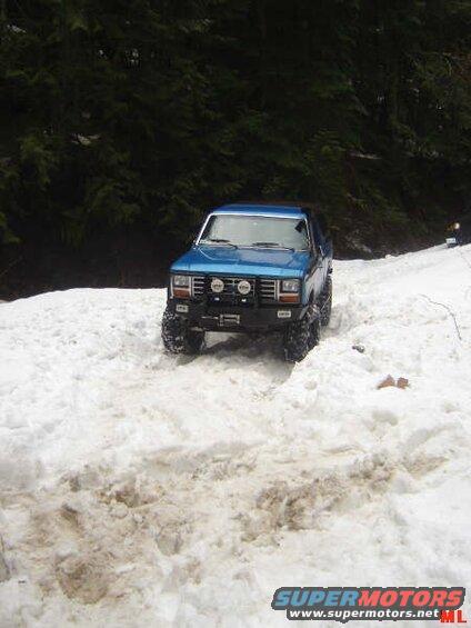 Blue 1980s Ford Bronco driving through snow on a wooded trail.
