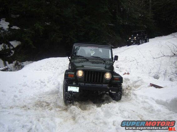 A dark green Jeep Wrangler driving on a snowy forest trail.